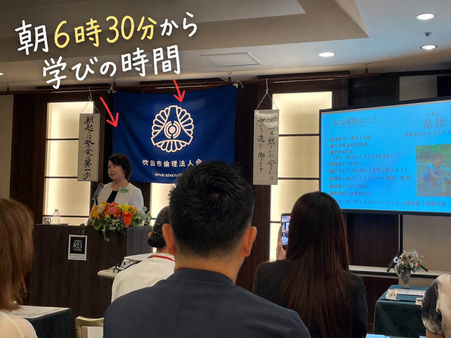 吹田市倫理法人会の会場。青い幕の前で島津郁代さんが講話をしている
